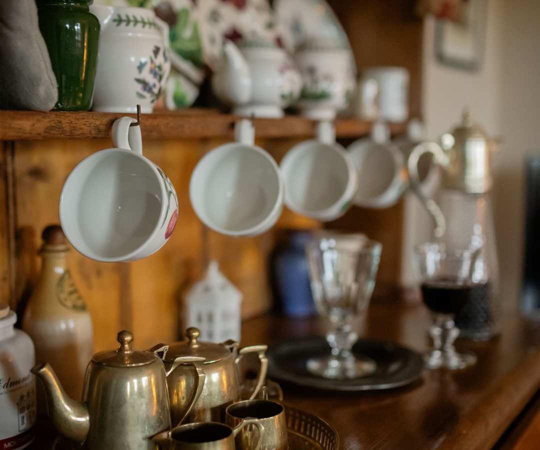 A wooden shelf in the cozy Gablebach nook holds hanging teacups and displayed plates, with brass teapots and cups on a surface below.