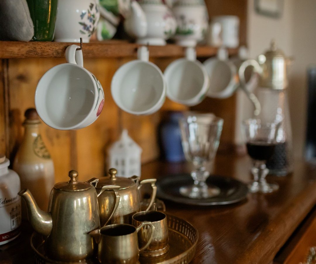 A wooden shelf in the cozy Gablebach nook holds hanging teacups and displayed plates, with brass teapots and cups on a surface below.