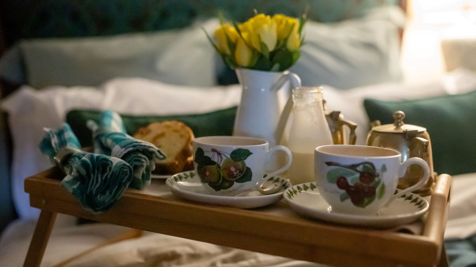 A breakfast tray on a bed at Gablebach with two teacups, a small milk jug, a loaf of bread, and a vase of yellow flowers—an ideal start to your serene Black Mountain Escapes.