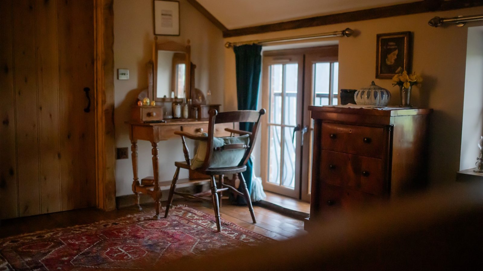 Cozy room with a wooden dresser, mirror, and chair near a window. A red patterned rug graces the wooden floor, complemented by warm lighting. Perfect for those who seek solace in mountain retreats or as a stop on your Black Mountain Escapes journey.