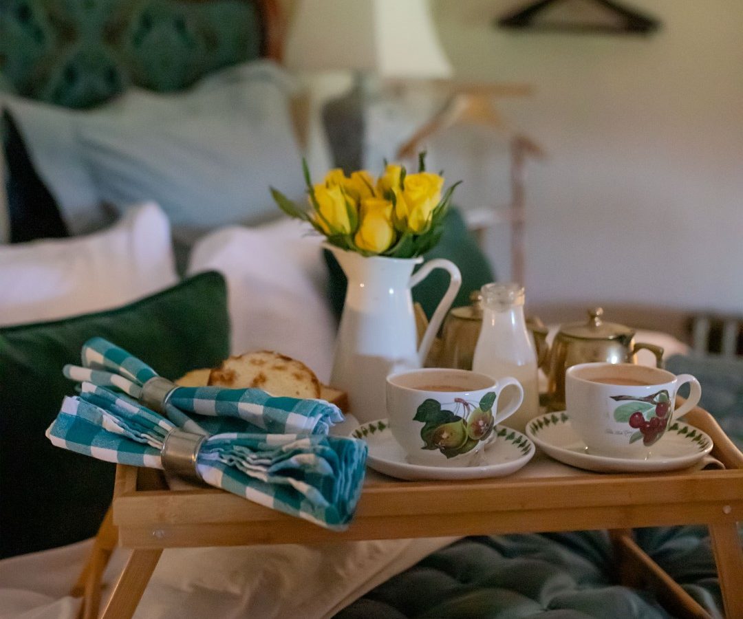 The breakfast tray, courtesy of Black Mountain Escapes, features two cups, a milk jug, napkins, buns, and a vase of yellow tulips perfectly arranged on a bed with green bedding.