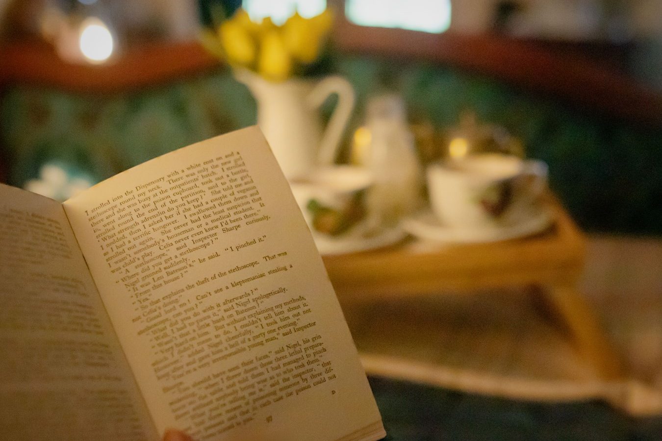 A person holds an open book, creating a cozy reading scene reminiscent of Black Mountain Escapes, with blurred cups and a jug on a tray in the background.