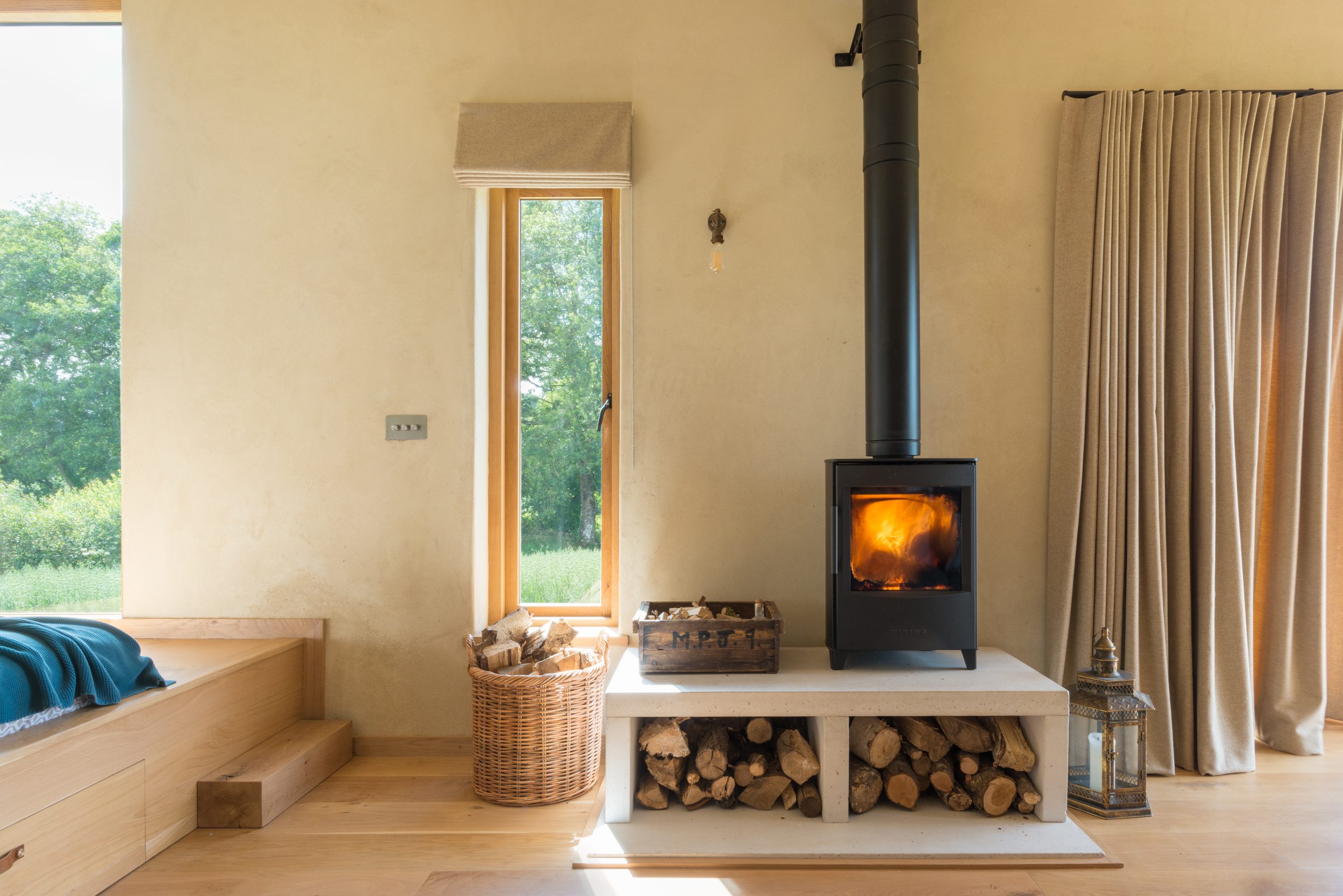 A wood stove with a fire burning, surrounded by firewood and a wicker basket, creates the perfect setting for cosy cabins for two. Nestled next to a window with a view of lush greenery, it invites relaxation and warmth.