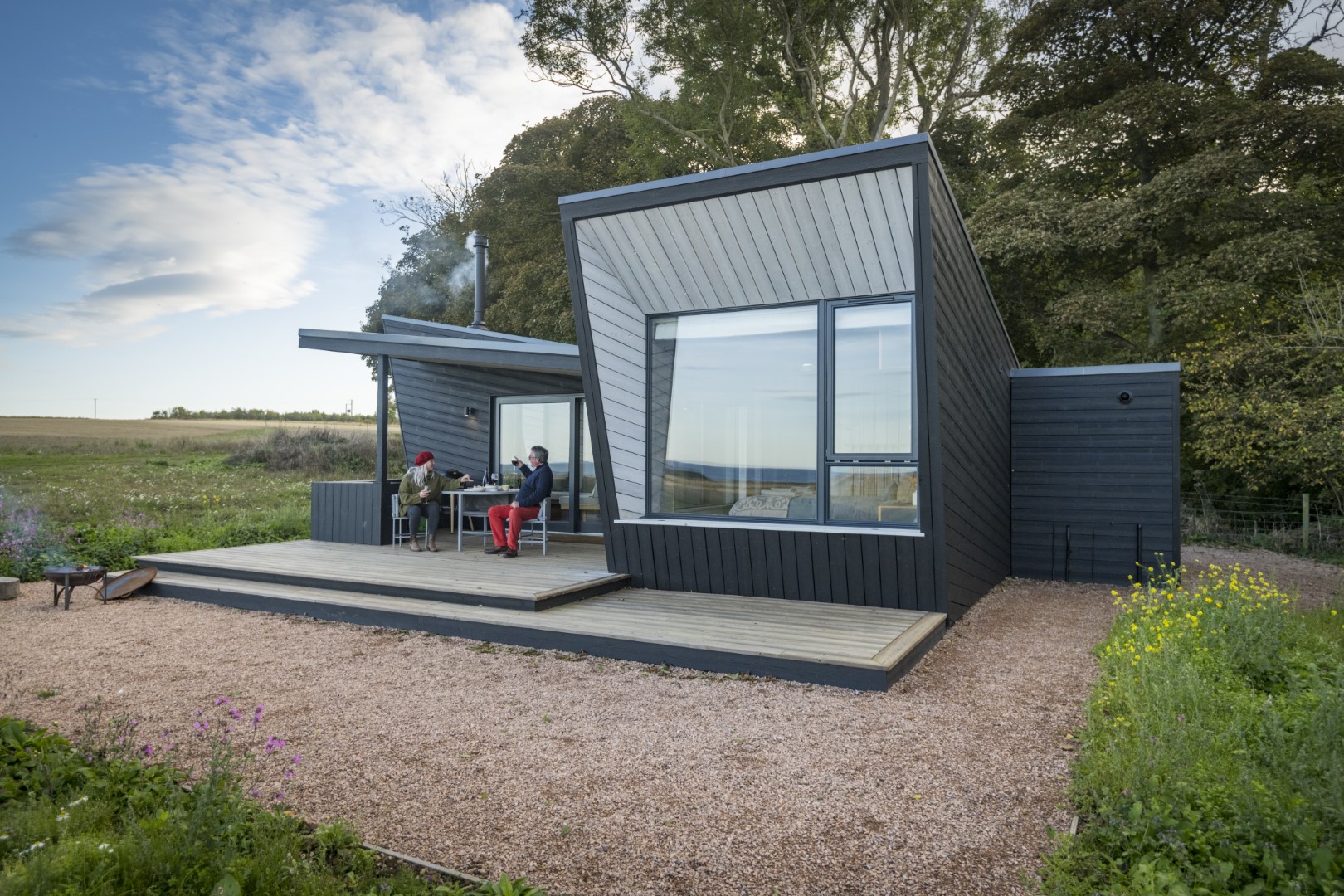 Modern black cabin with large windows and a wooden deck, surrounded by greenery; perfect for holiday lets through local partnerships. Two people sit outside on the deck, enjoying the serene environment.