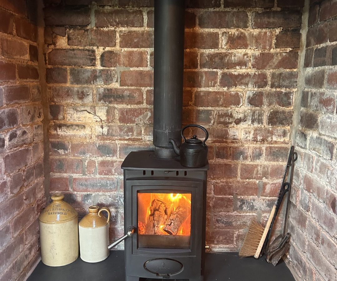 A cozy scene with a wood-burning stove at Black Mountain Escapes, featuring a kettle on top and encircled by brick walls. Nearby, two jugs, tongs, and a brush complete the rustic Y Golchdy charm.