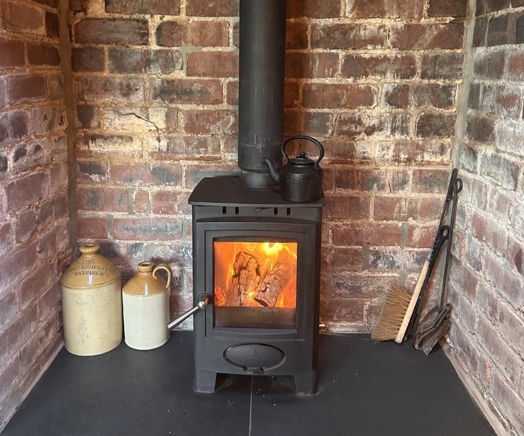 A cozy scene with a wood-burning stove at Black Mountain Escapes, featuring a kettle on top and encircled by brick walls. Nearby, two jugs, tongs, and a brush complete the rustic Y Golchdy charm.