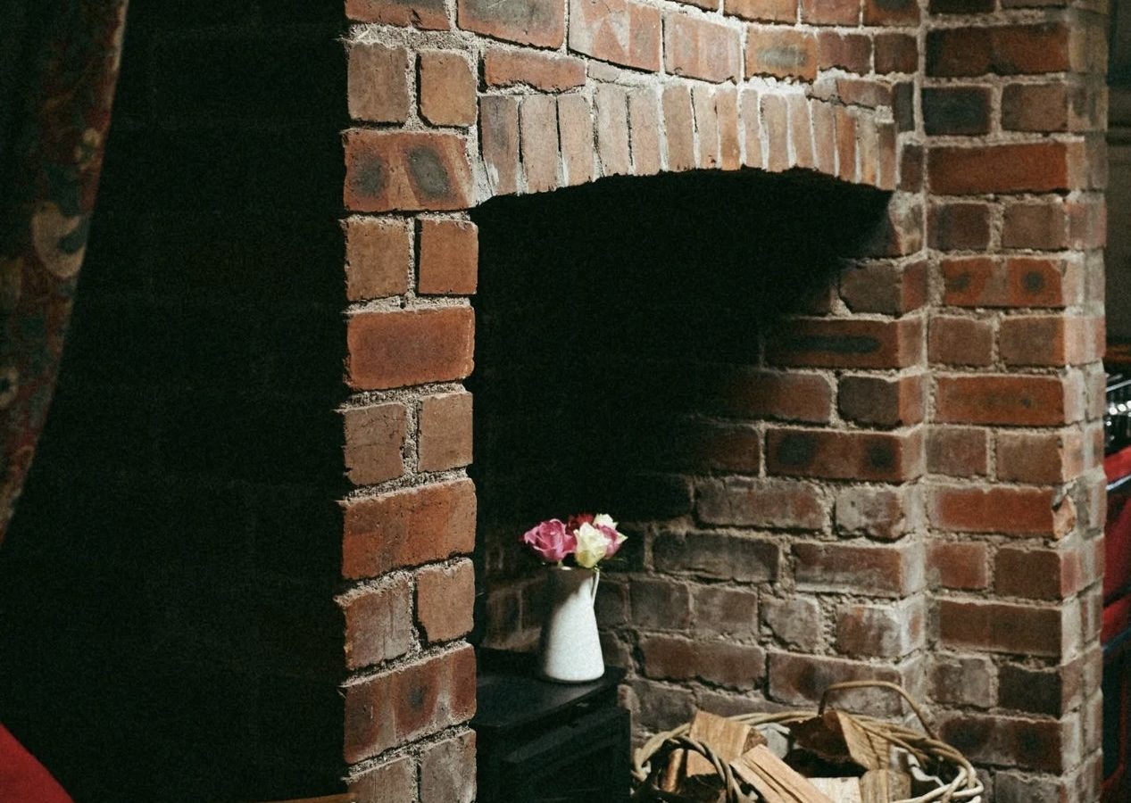 A cozy corner by the brick fireplace features a black wood stove, a woven basket of firewood, and a small vase with flowers gracing the mantle. This inviting setting is reminiscent of charming retreats offered by Black Mountain Escapes.