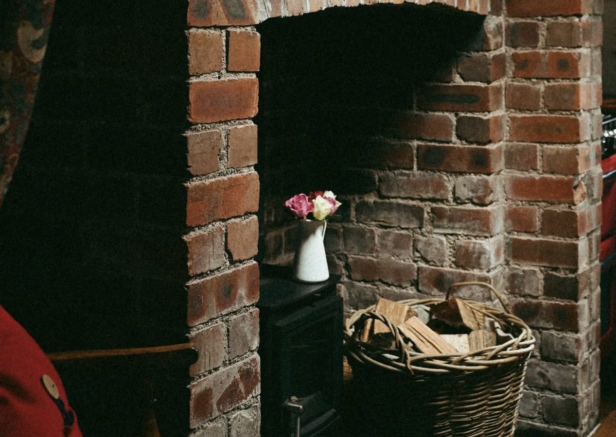 A cozy corner by the brick fireplace features a black wood stove, a woven basket of firewood, and a small vase with flowers gracing the mantle. This inviting setting is reminiscent of charming retreats offered by Black Mountain Escapes.