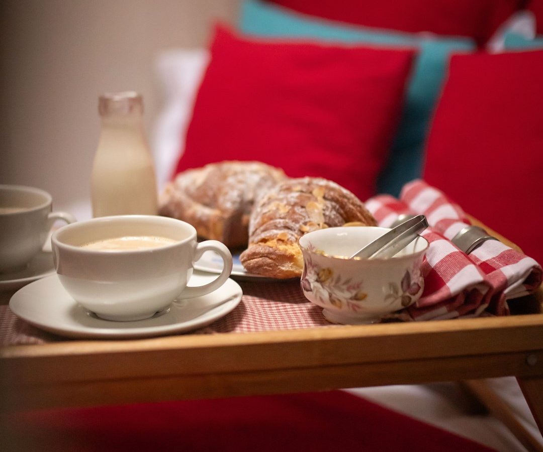 A breakfast tray featuring coffee, croissants, and a milk bottle sits elegantly with cutlery on a bed adorned with red and teal pillows at Black Mountain Escapes.