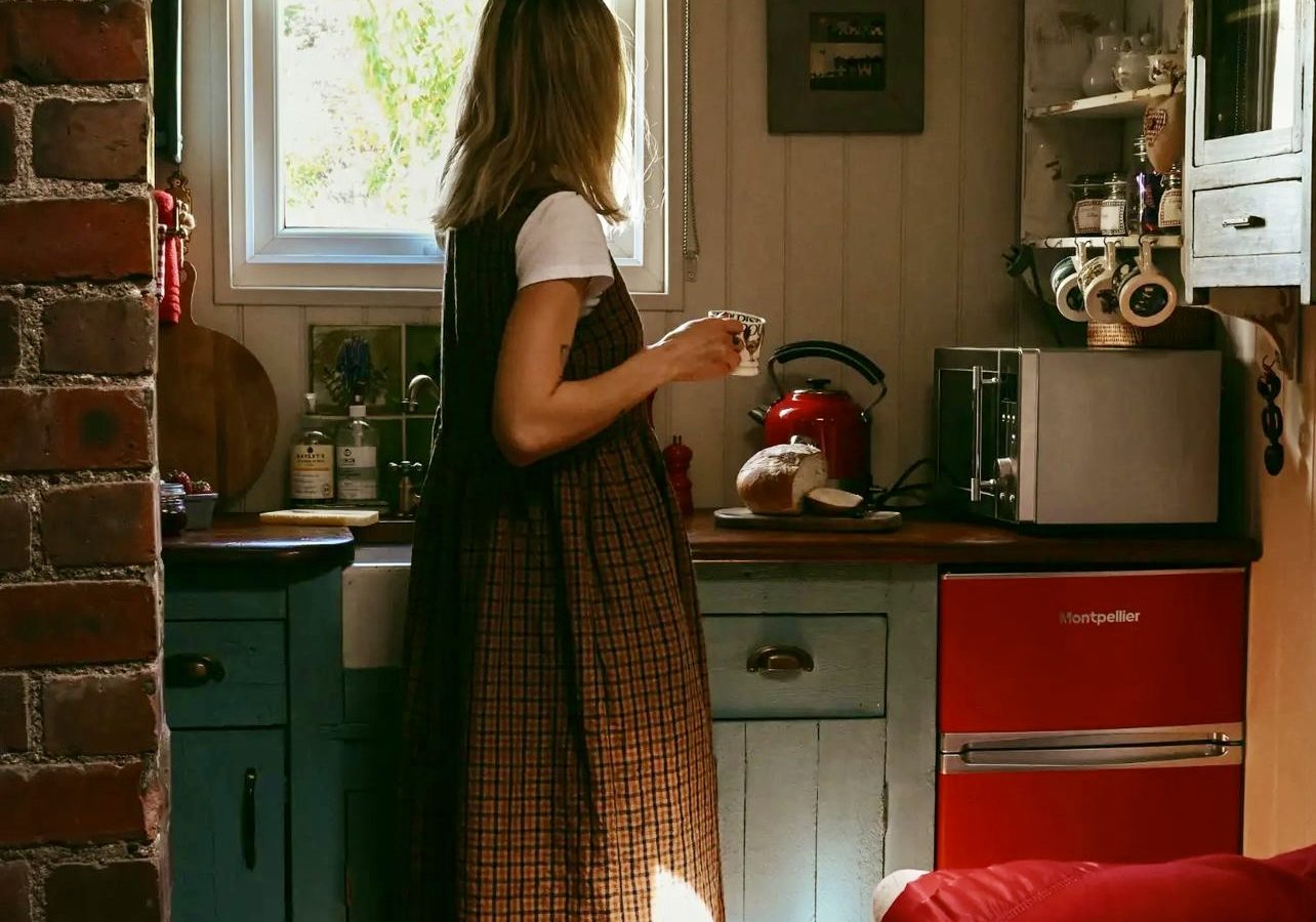 A woman holds a mug, gazing out a small kitchen window adorned with vintage decor, wooden cabinets, and a small red refrigerator—a cozy scene reminiscent of Black Mountain Escapes.