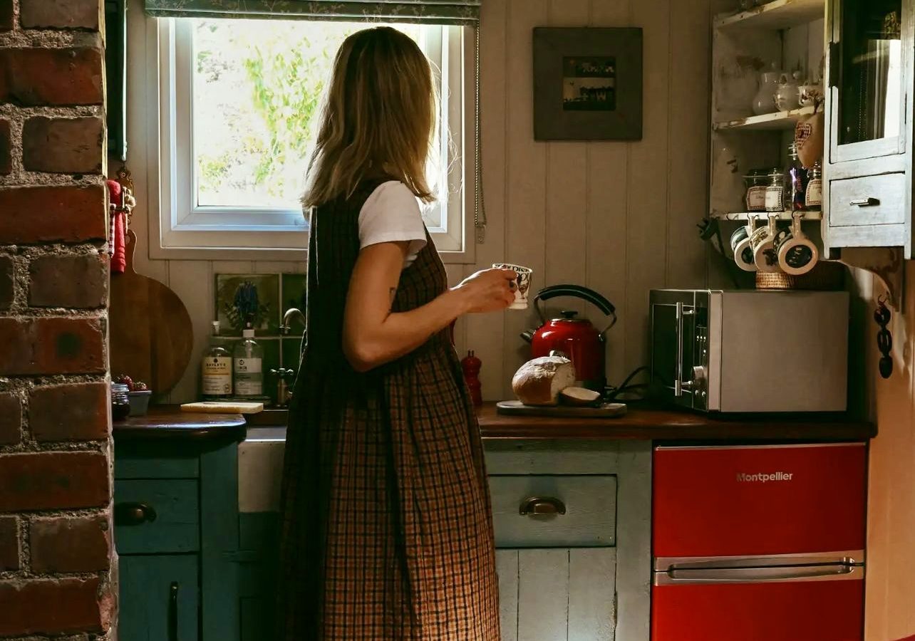 A woman holds a mug, gazing out a small kitchen window adorned with vintage decor, wooden cabinets, and a small red refrigerator—a cozy scene reminiscent of Black Mountain Escapes.