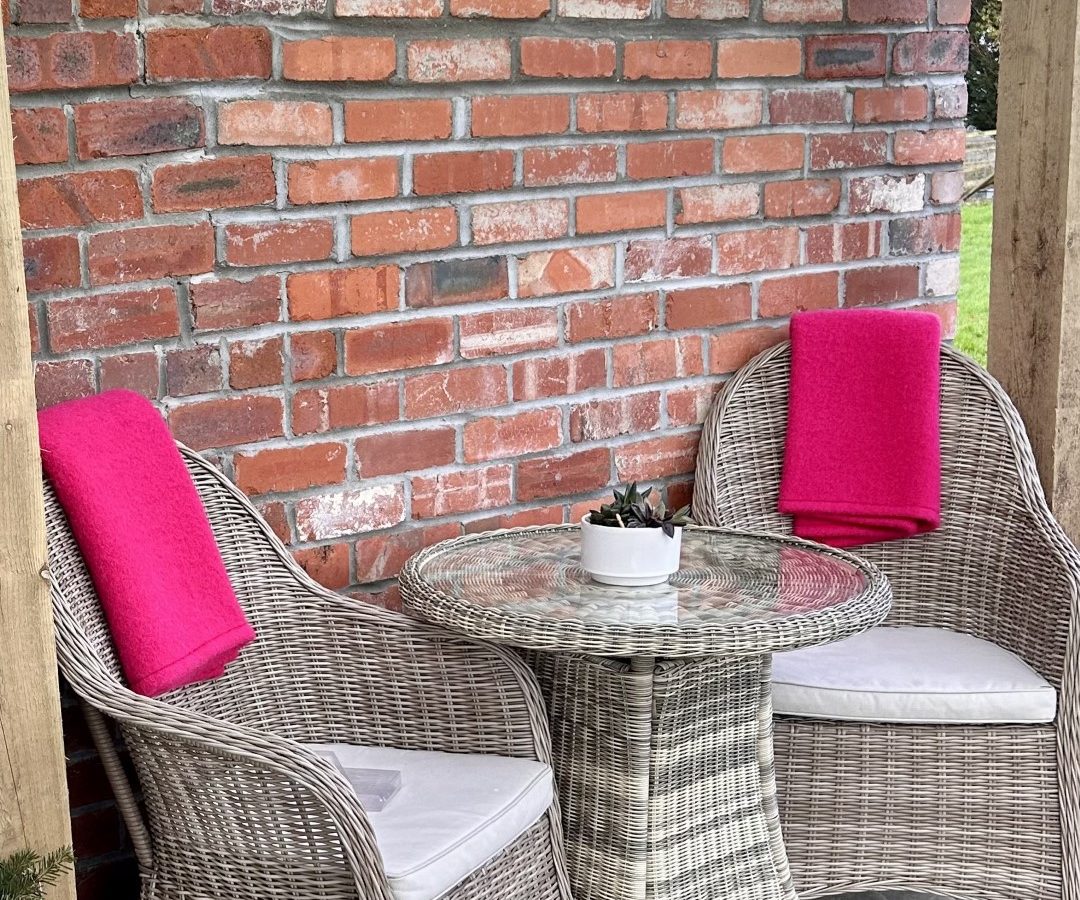 Two wicker chairs with pink cushions and a round glass table grace the stone patio, nestled against a red brick wall. This serene setting is perfect for unwinding at Y Golchdy, part of the Black Mountain Escapes collection.