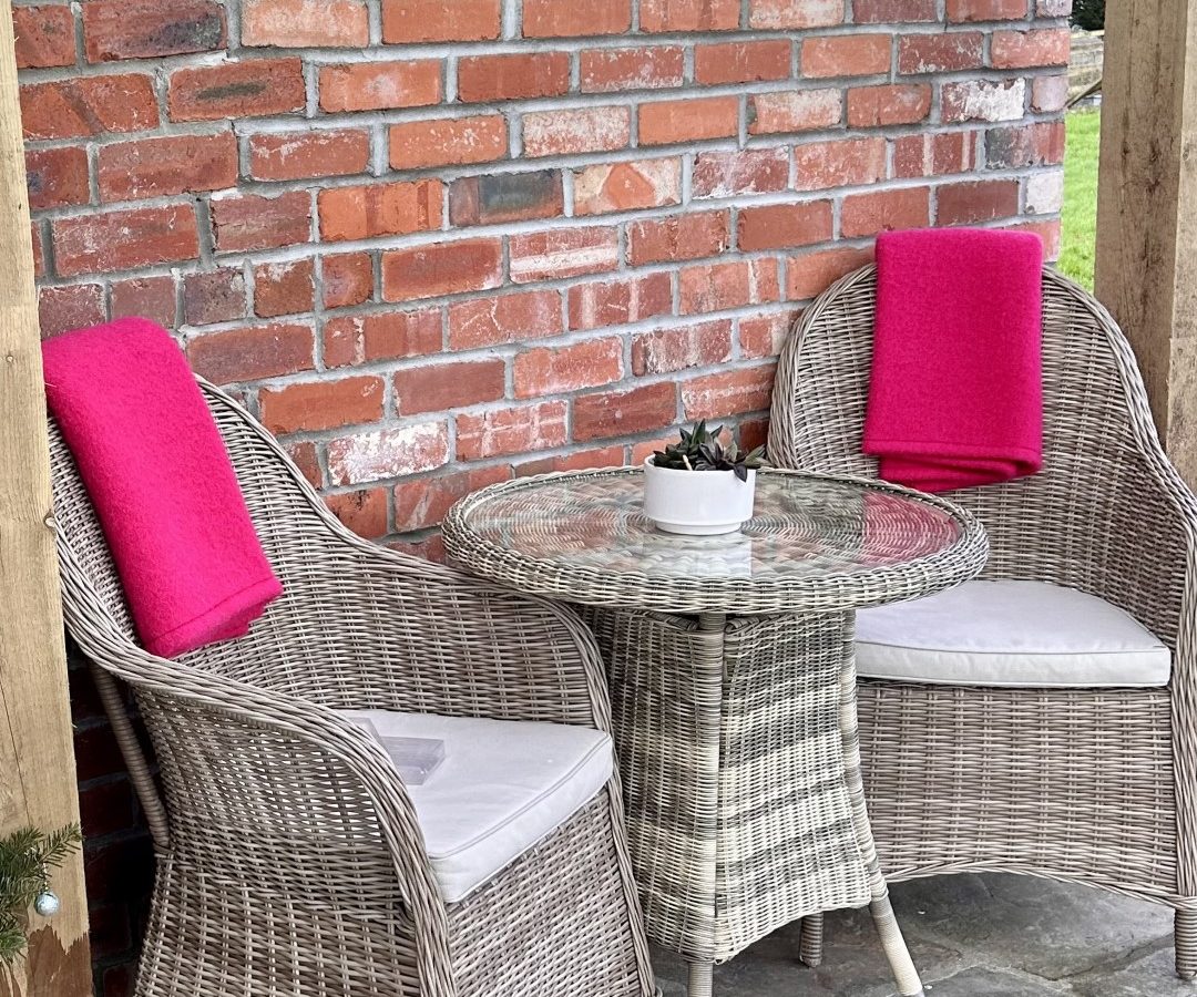 Two wicker chairs with pink cushions and a round glass table grace the stone patio, nestled against a red brick wall. This serene setting is perfect for unwinding at Y Golchdy, part of the Black Mountain Escapes collection.