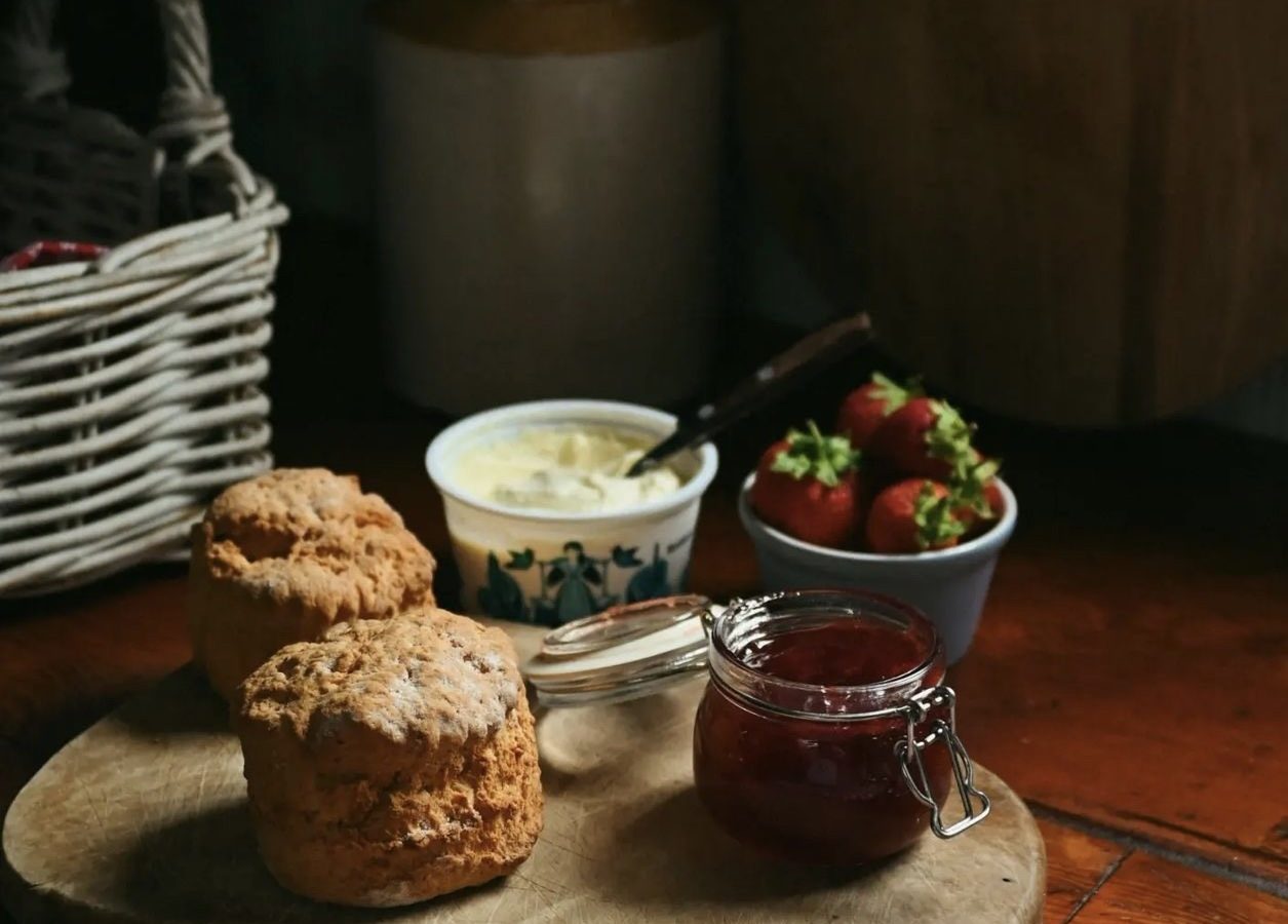 Scones on a wooden board with a jar of jam, a bowl of strawberries, and a pot of cream create a cozy scene at Black Mountain Escapes, set on the rustic kitchen table of Y Golchdy.