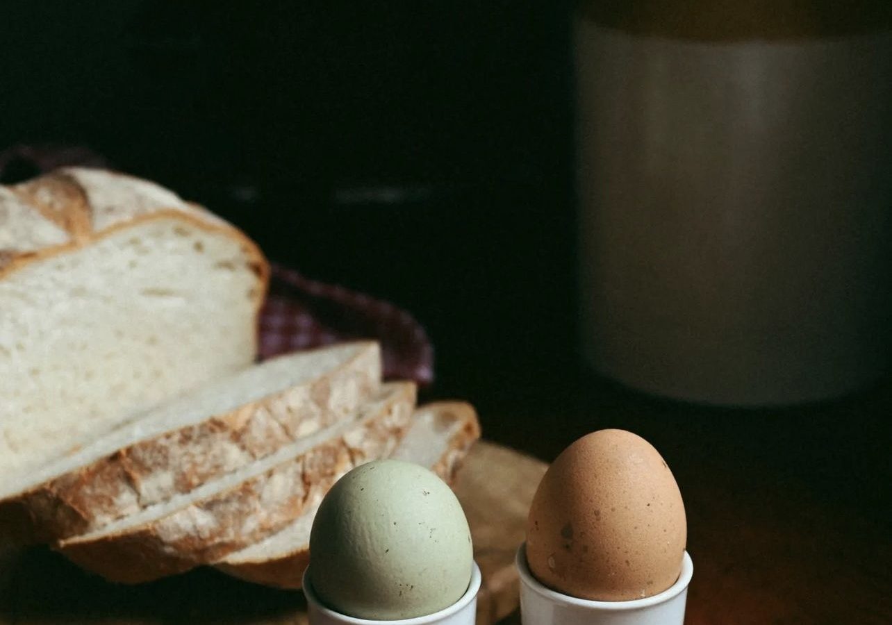 Two eggs in white holders sit on a wooden table near sliced bread, evoking the rustic charm of Black Mountain Escapes. In the background, a jar containing utensils hints at the inviting ambiance of Y Golchdy.