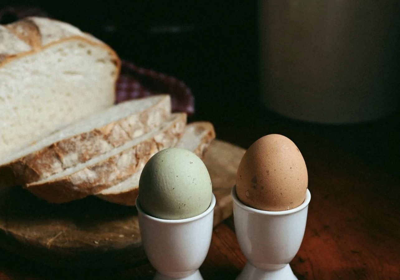 Two eggs in white holders sit on a wooden table near sliced bread, evoking the rustic charm of Black Mountain Escapes. In the background, a jar containing utensils hints at the inviting ambiance of Y Golchdy.