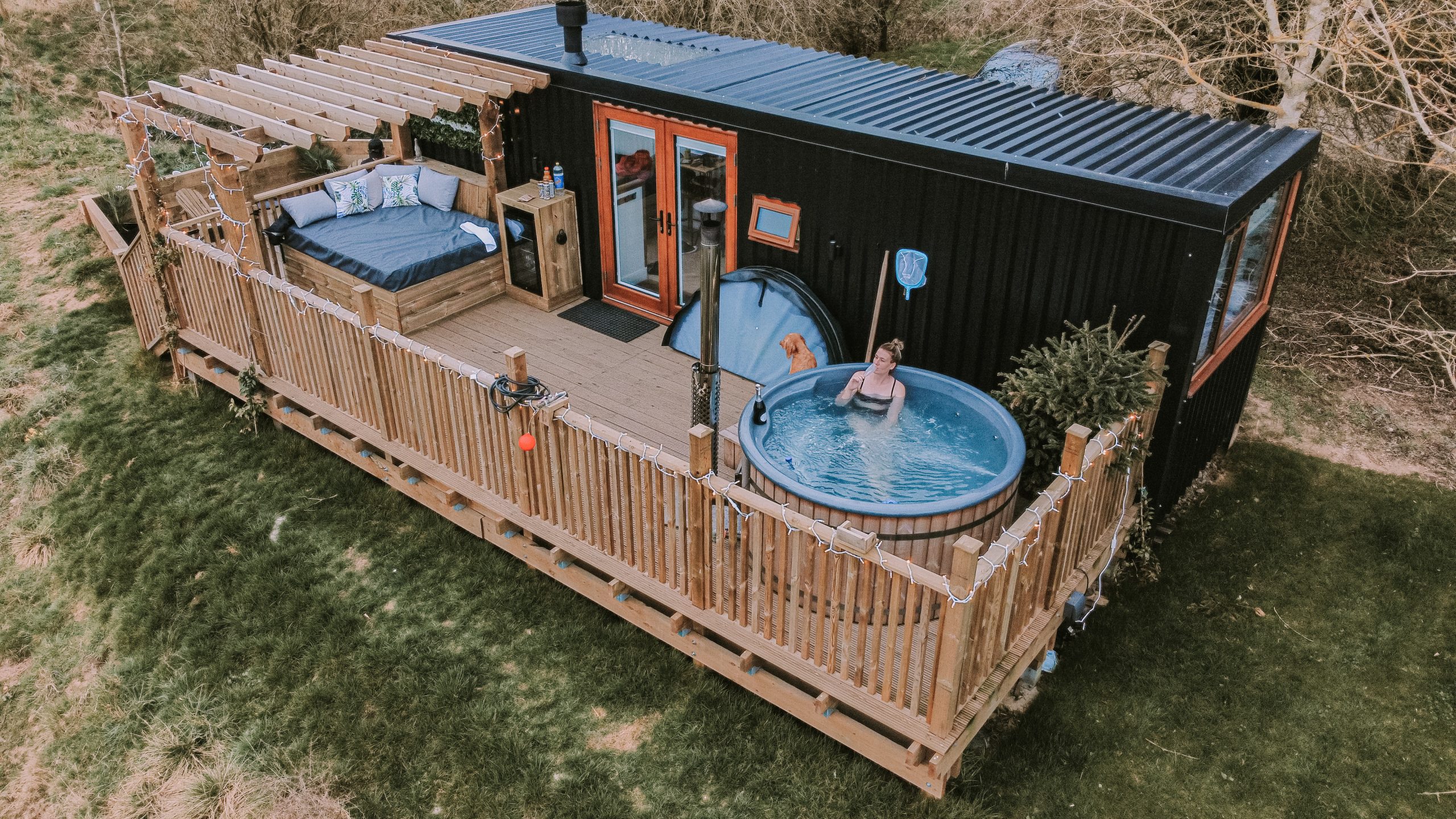 Aerial view of a black shipping container home featuring a wooden deck, hot tub, and seating area. This cosy cabin for two is surrounded by lush greenery, offering the perfect escape into nature.