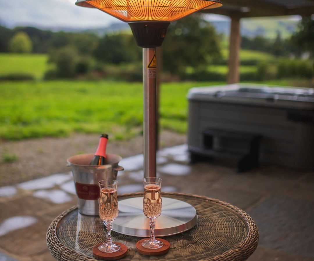 On the Golchdy terrace, a wicker table hosts two champagne glasses and a bottle in a cooler, all warmed by a patio heater. The serene setting is part of the Black Mountain Escapes collection, offering tranquil views over lush green fields.