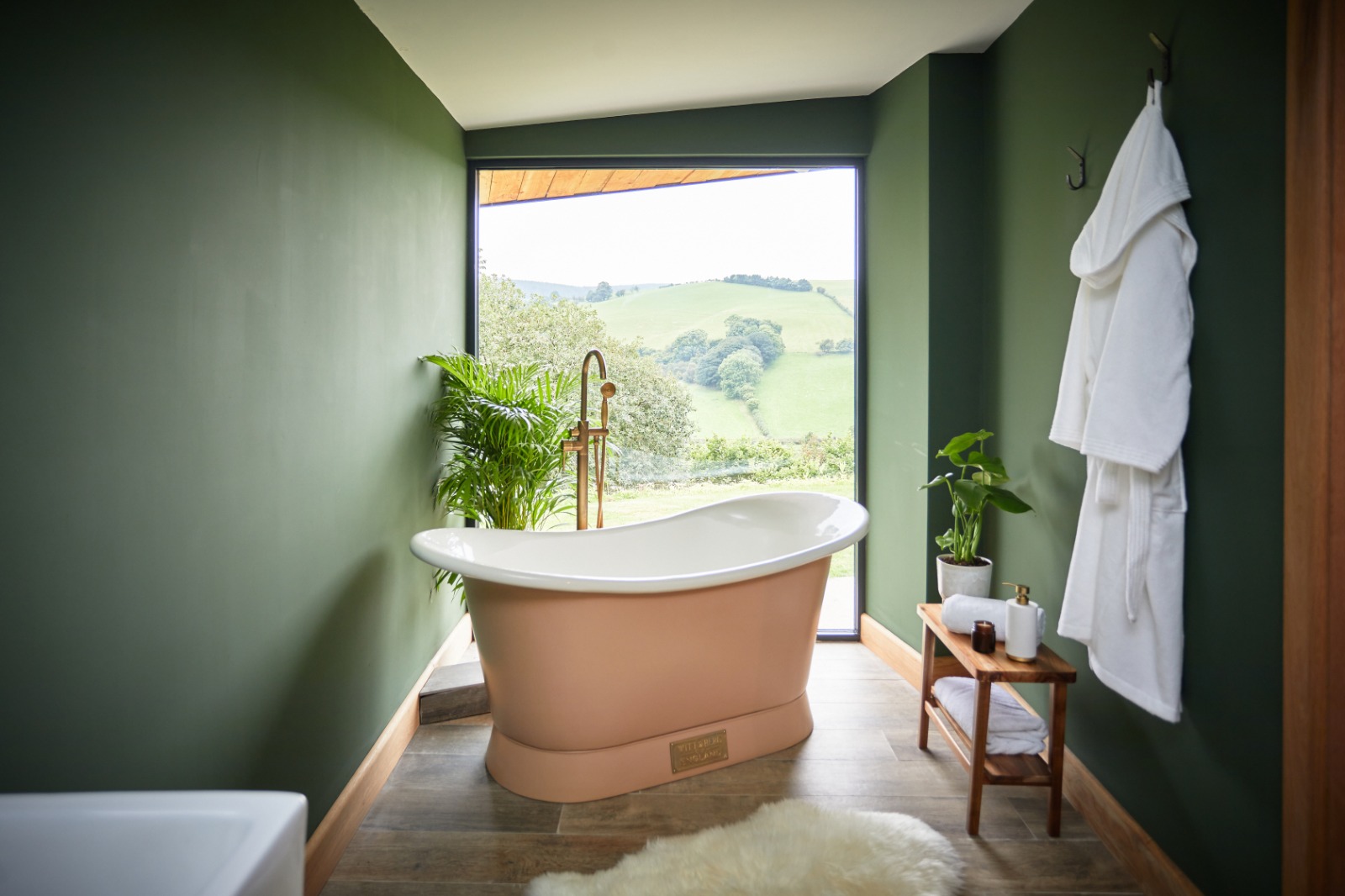 Experience design-led stays in this green-walled bathroom featuring a freestanding bathtub, where you can soak while enjoying the large window's view of rolling hills. Complemented by a fresh plant and towel on a rack, it's an oasis of relaxation.