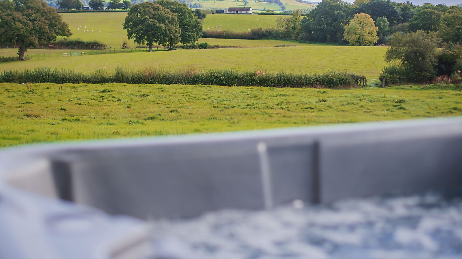 A bubbling hot tub is in the foreground, overlooking a lush, green countryside with rolling hills under a cloudy sky. Trees and hedgerows dot the landscape, creating a serene and inviting rural scene at Black Mountain Escapes.