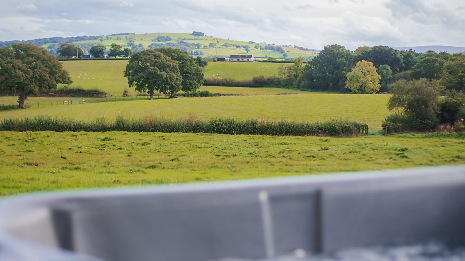 A bubbling hot tub is in the foreground, overlooking a lush, green countryside with rolling hills under a cloudy sky. Trees and hedgerows dot the landscape, creating a serene and inviting rural scene at Black Mountain Escapes.