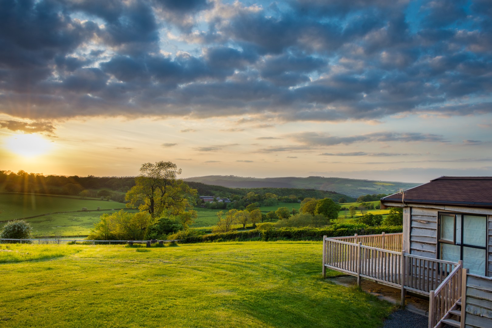 This Shropshire destination features a house with a deck that overlooks a grassy field at sunset, framed by trees and hills under a partly cloudy sky—truly a must-visit for nature lovers.