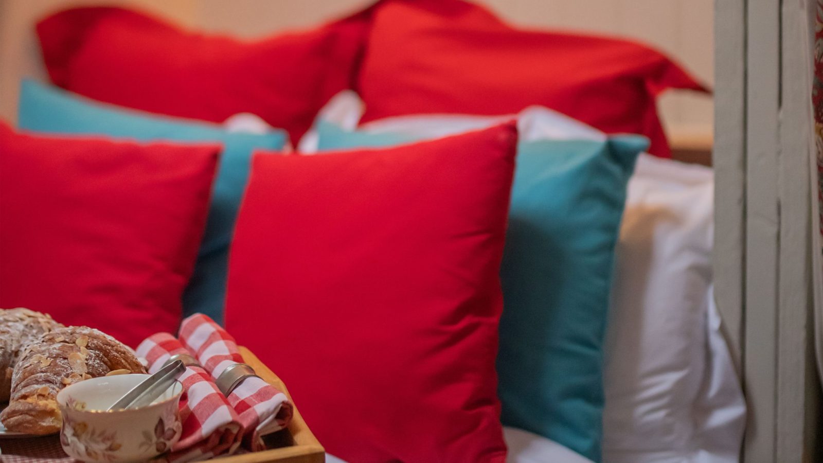 A cozy bedroom at Black Mountain Escapes features a bed with red and turquoise pillows. A wooden tray holds breakfast rolls and a cup, while warm lighting from two wall-mounted lamps above creates a soft, inviting ambiance reminiscent of Y Golchdy’s charm.