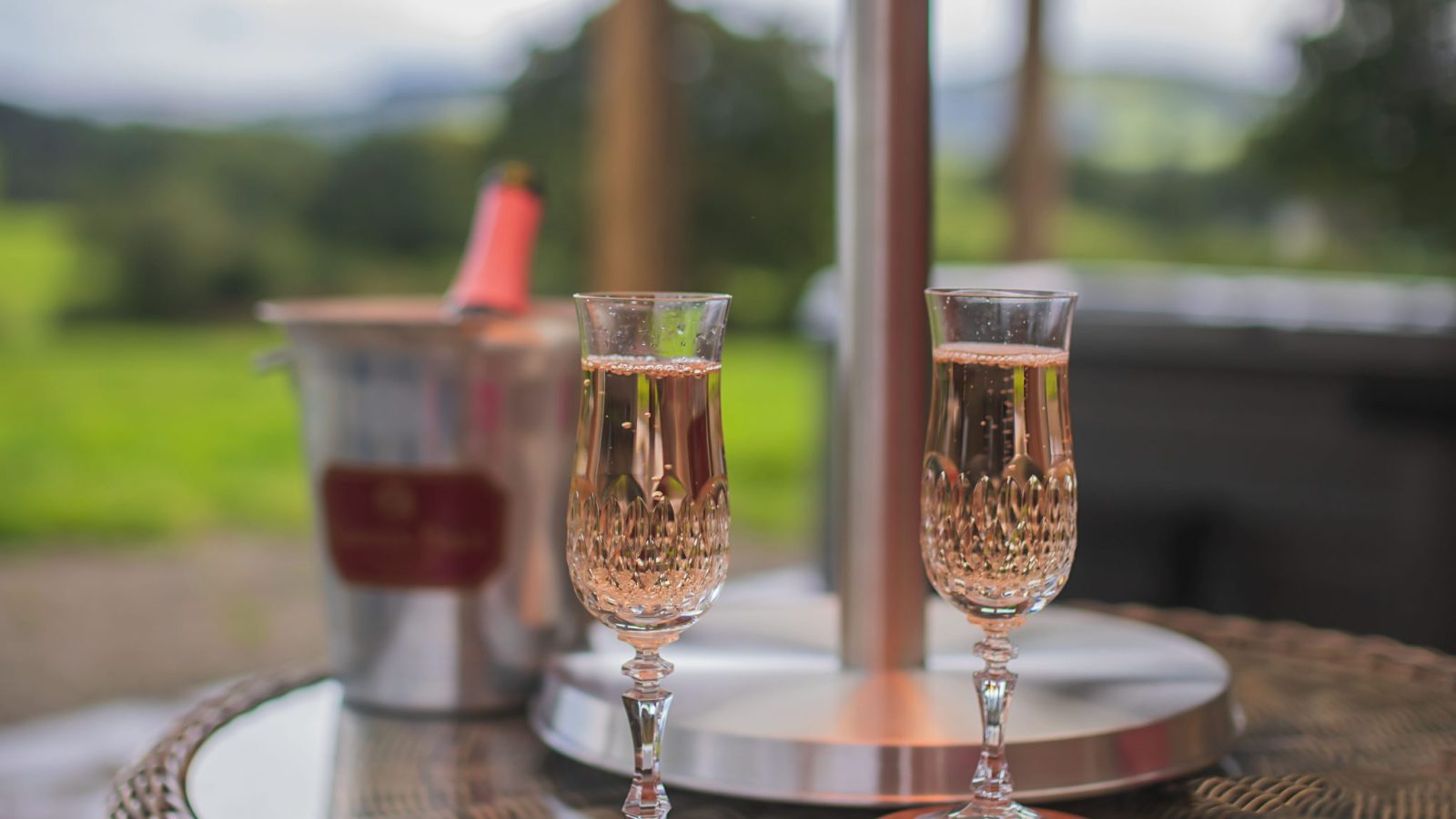 Two crystal champagne flutes rest on a patio table beneath a heater at Black Mountain Escapes, overlooking a lush landscape. A chilled bottle waits in a metal ice bucket nearby, creating the perfect Golchdy retreat.