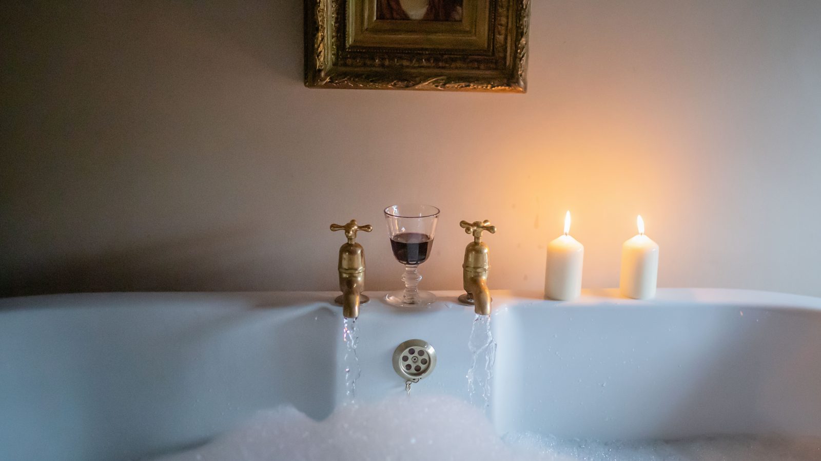 A filled bathtub with soap bubbles offers a perfect Black Mountain escape. A glass of red wine rests on the ledge, while two lit candles create a warm glow. An ornate framed portrait hangs above, adding a cozy ambiance reminiscent of Gablebach charm.