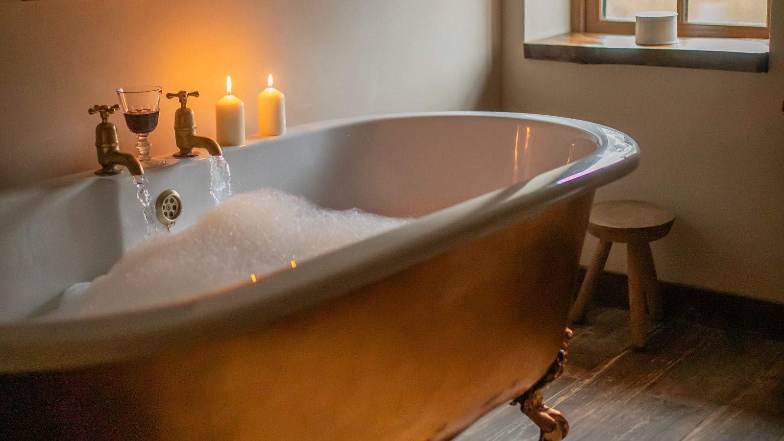 A cozy bathroom by Black Mountain features a vintage-style freestanding bathtub filled with bubbles. Lit candles rest on the edge, creating a warm ambiance. A small window above provides soft natural light, and a painting hangs on the wall nearby, offering perfect escapes from daily life.