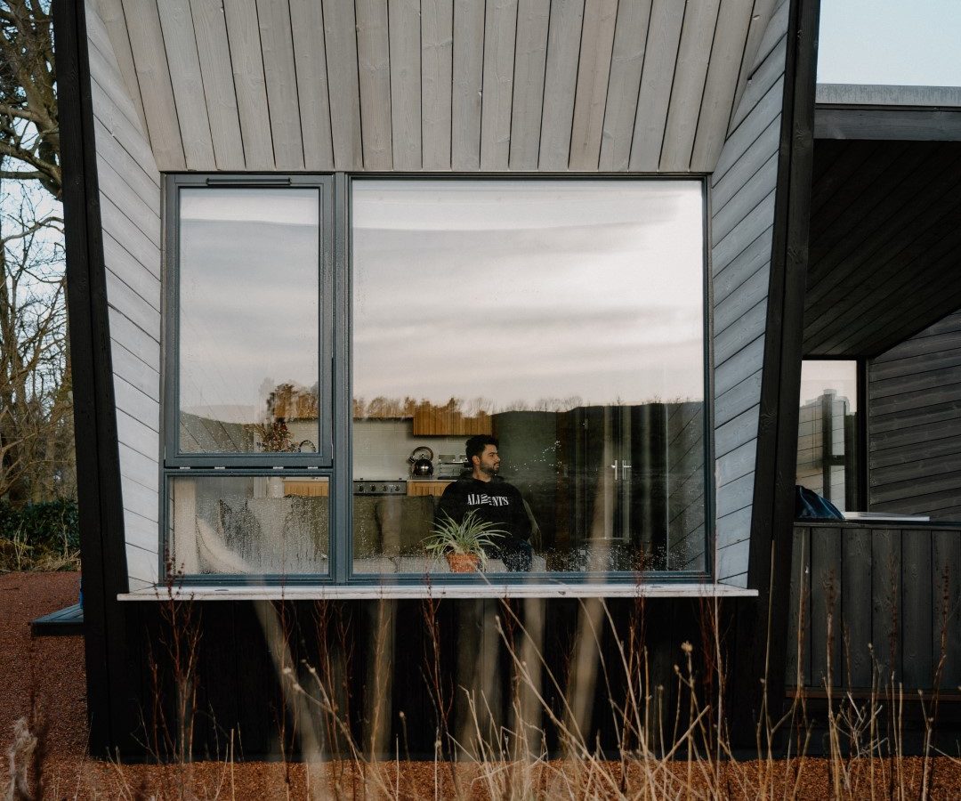 A modern wooden house on the Cambo Estate features large glass windows mirroring the sky and surrounding nature. A person is visible inside, seated serenely. The house, perfect for off-grid travel enthusiasts, is embraced by tall grasses and trees beneath a clear blue sky.