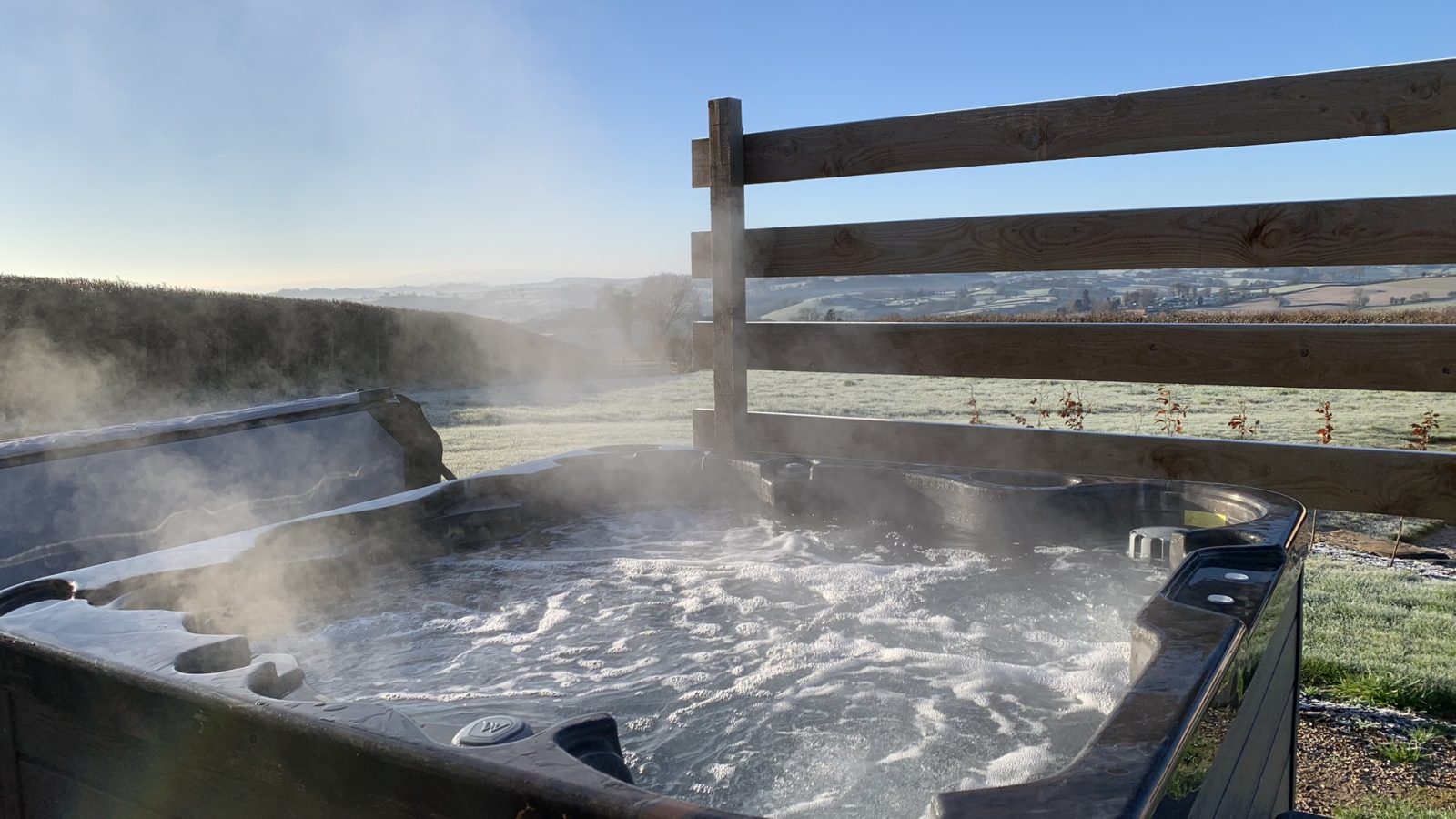 A steaming hot tub outdoors at Belan View lodges, surrounded by a wooden fence, overlooks a scenic landscape with frost-covered fields and hills under a clear blue sky.