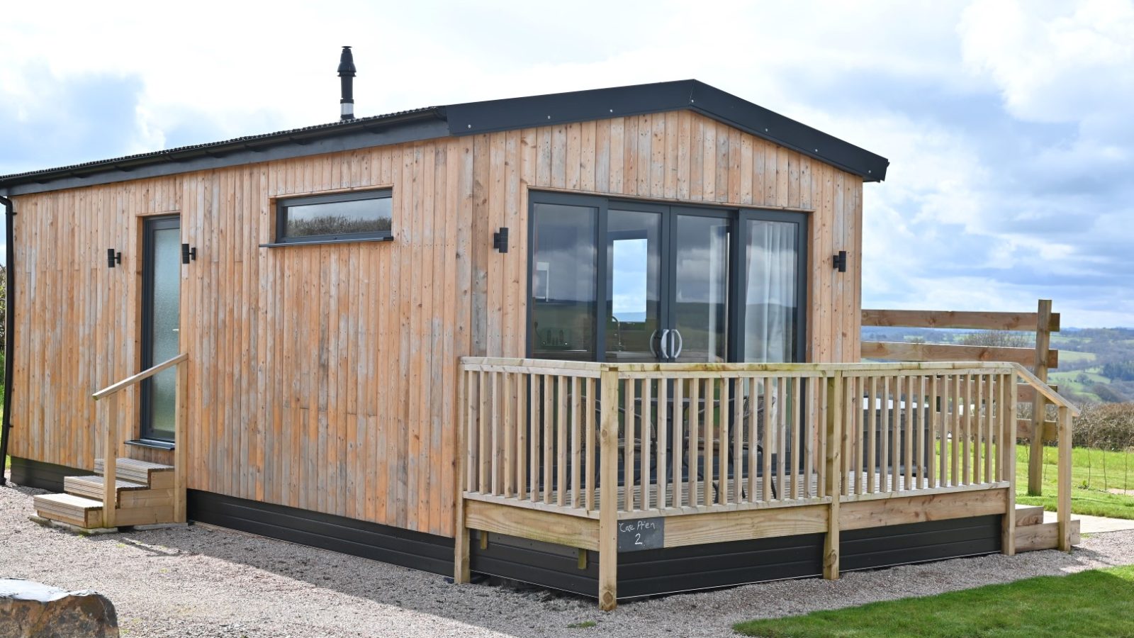 Belan View is a small, modern wooden cabin with large windows and a porch, nestled on a gravel path. It overlooks a vast, green landscape under a partly cloudy sky, offering serene beauty.