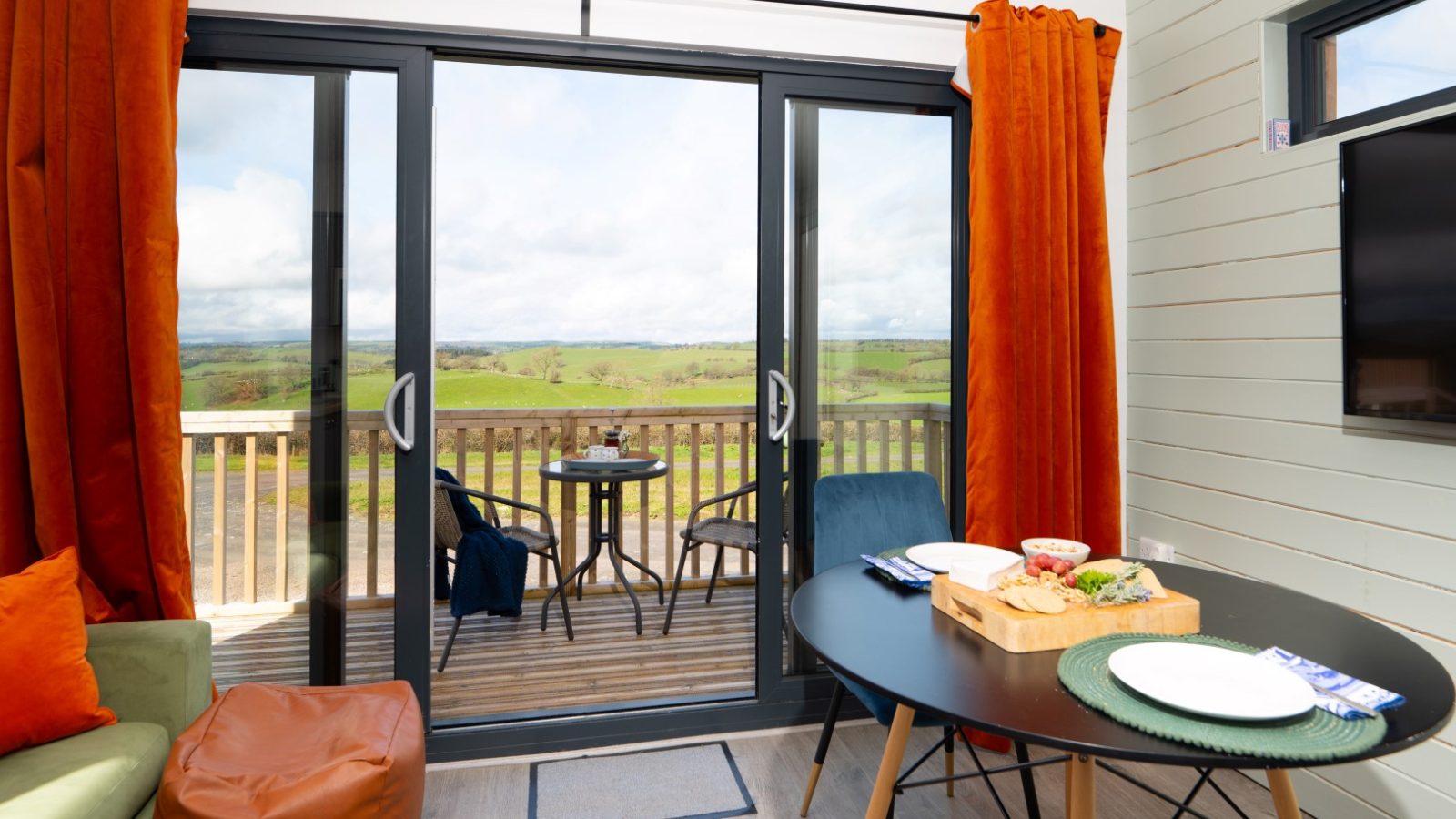 The cozy living room at Belan View features vibrant orange curtains and a glass door opening to a wooden deck. The deck, perfect for those staying at the lodges, has a small table with chairs offering a view of the vast green countryside under a cloudy sky. A round dining table with plates is indoors.