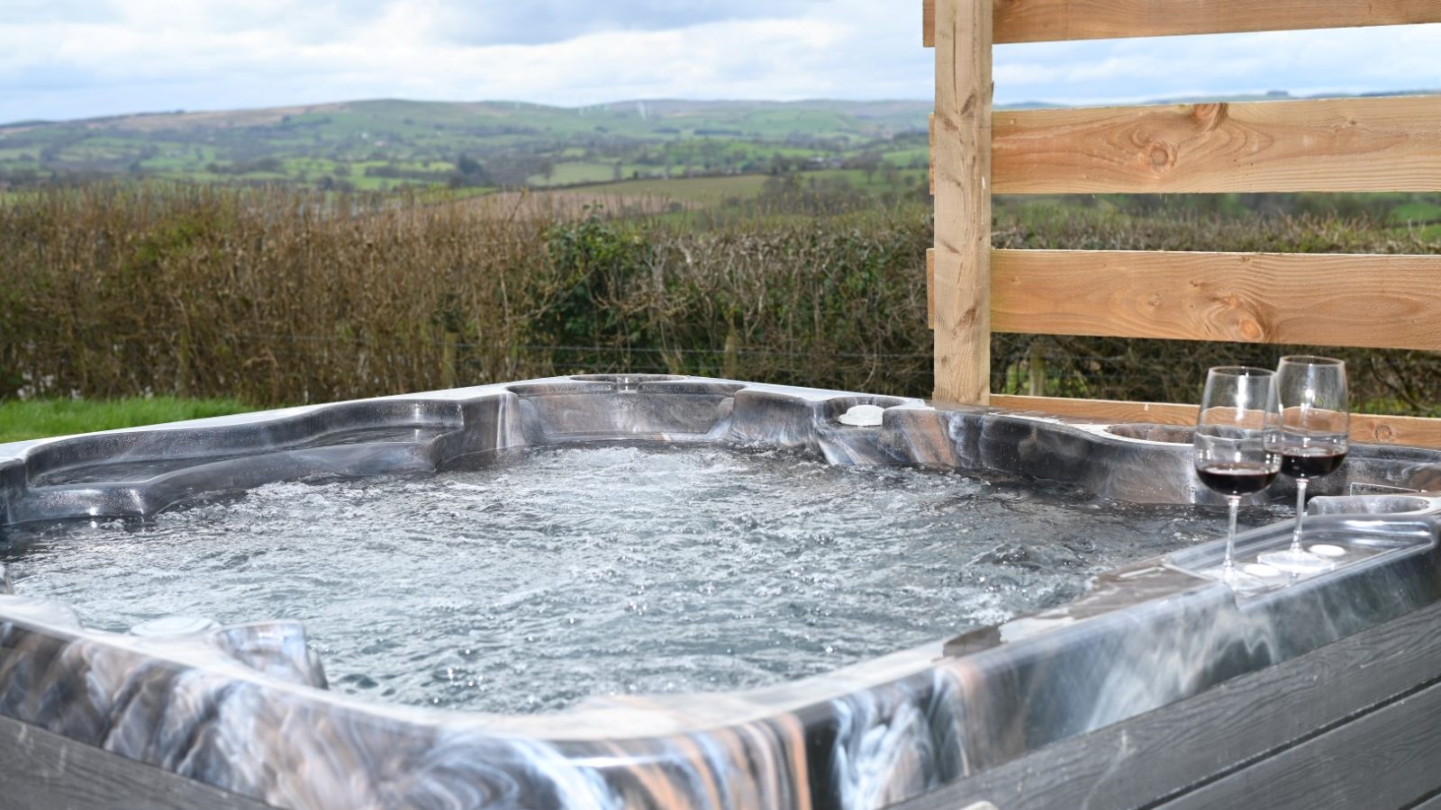 A bubbling hot tub on a wooden deck at Belan View overlooks the scenic countryside with rolling hills. Two glasses of red wine rest nearby, and a wooden privacy screen adds seclusion to this idyllic lodge experience. The sky is cloudy yet bright, enhancing the serene atmosphere.