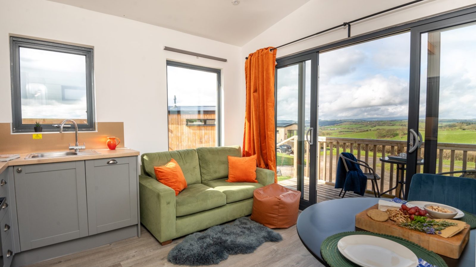 A cozy living area with a green sofa and orange cushions, nestled in Belan View Lodges, adjacent to a kitchen with sleek gray cabinets. Large sliding glass doors open to a wooden deck with countryside views. The dining table features dishes and a cutting board with assorted food.