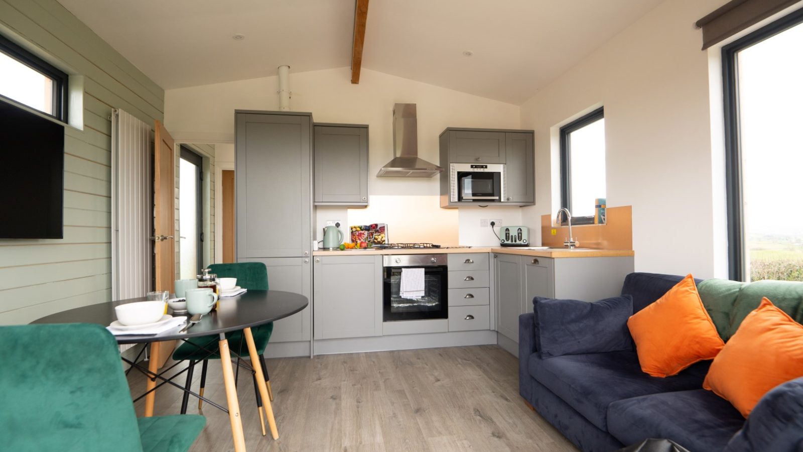 Nestled in Belan View, this modern, compact kitchen and living area boasts gray cabinets, a stainless steel oven and microwave. It features a seating area with a dark sofa, orange and green cushions, a round black dining table with green chairs, and large windows that flood the space with light.