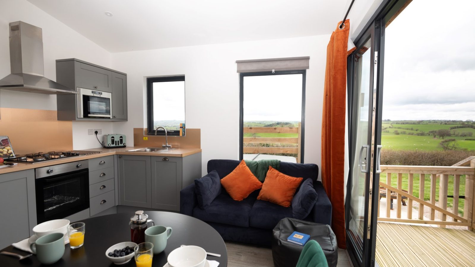 Modern kitchen and living area with gray cabinets, a round dining table set, and a dark sofa with orange cushions. Large sliding glass doors open to a wooden deck overlooking the scenic countryside, offering the perfect retreat at Belan View Lodges.