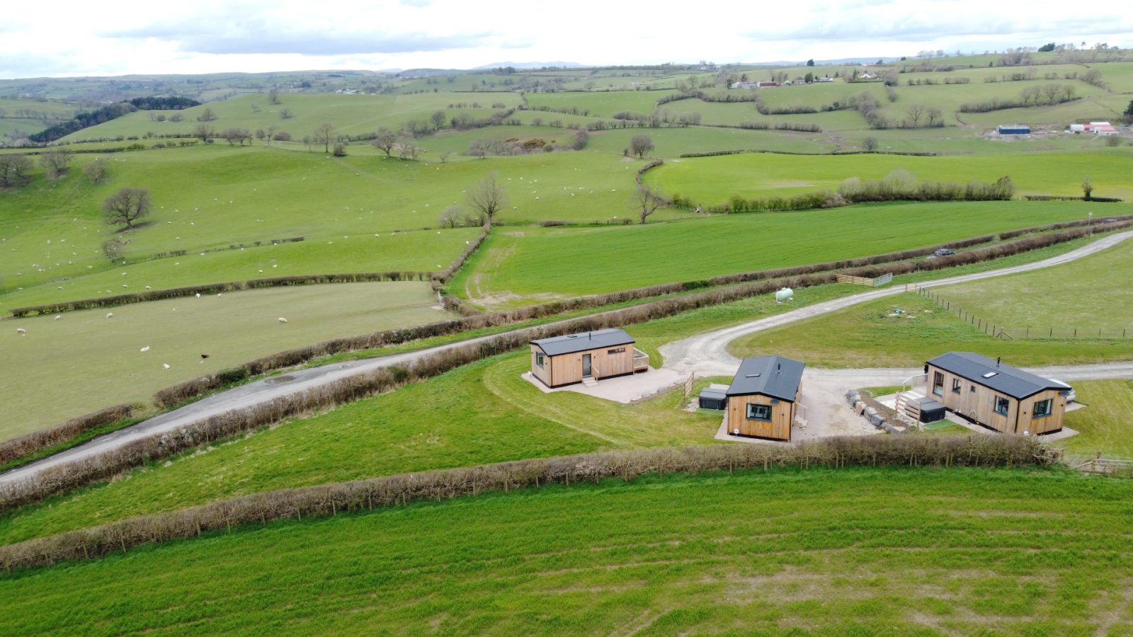 Aerial view of a rural landscape with rolling green hills. Three small, modern wooden lodges are scattered among fields and hedgerows at Belan View. A winding dirt road connects the lodges. Sheep graze in the distance under a cloudy sky.