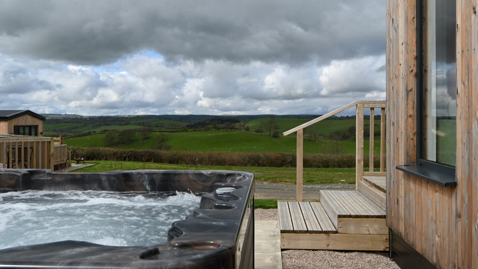 A hot tub on a wooden deck at Belan View overlooks a grassy landscape with rolling hills and cloudy skies. A set of steps leads to The Lodges, a modern wooden building on the right.