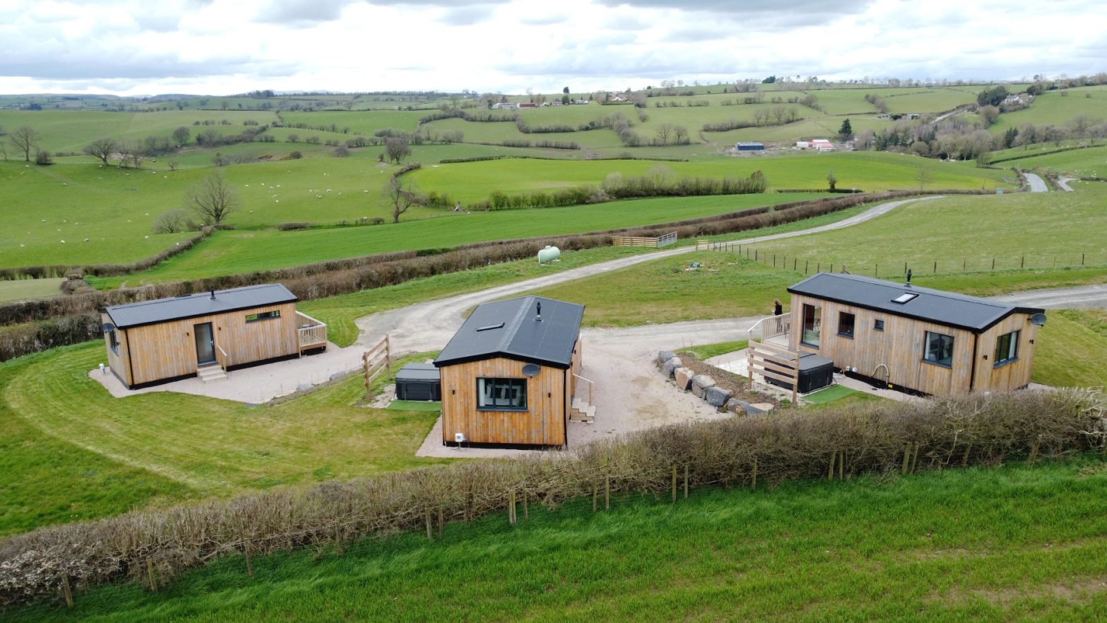 Nestled in a lush green countryside, The Lodges offer a scenic view of three small wooden cabins surrounded by fields and trees under a cloudy sky. A dirt path connects the cabins at Belan View, with rolling hills gracing the background.