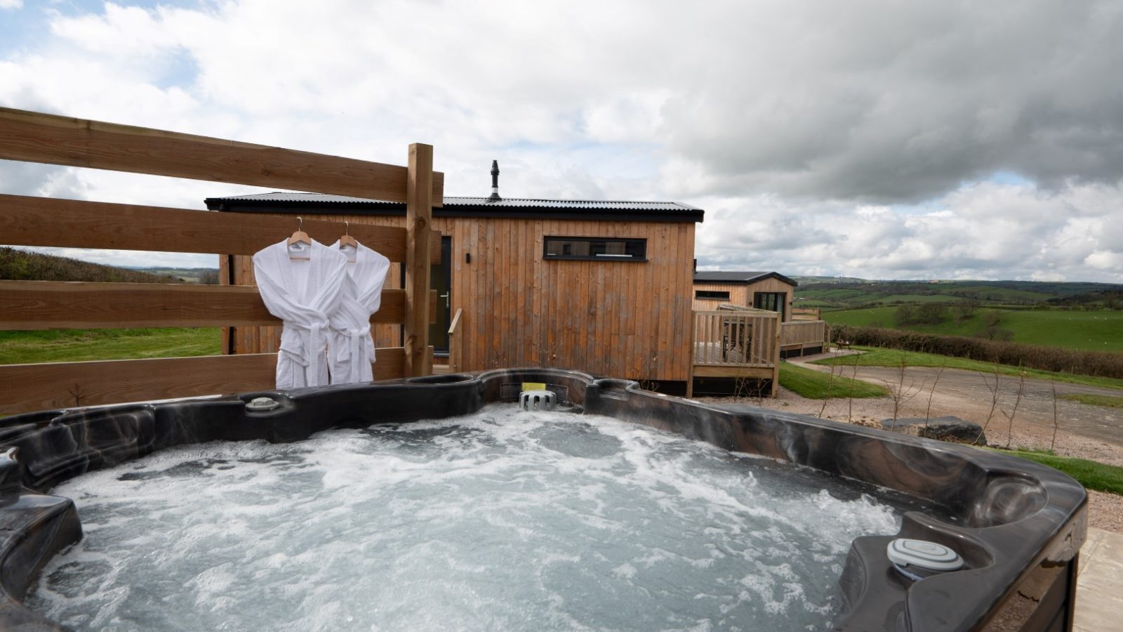 Belan View offers a charming wooden cabin, complete with a hot tub nestled in the foreground, surrounded by a stunning rural landscape. Two white bathrobes hang on the fence, enhancing the cozy atmosphere beneath the cloudy sky and revealing scenic green fields and distant hills.