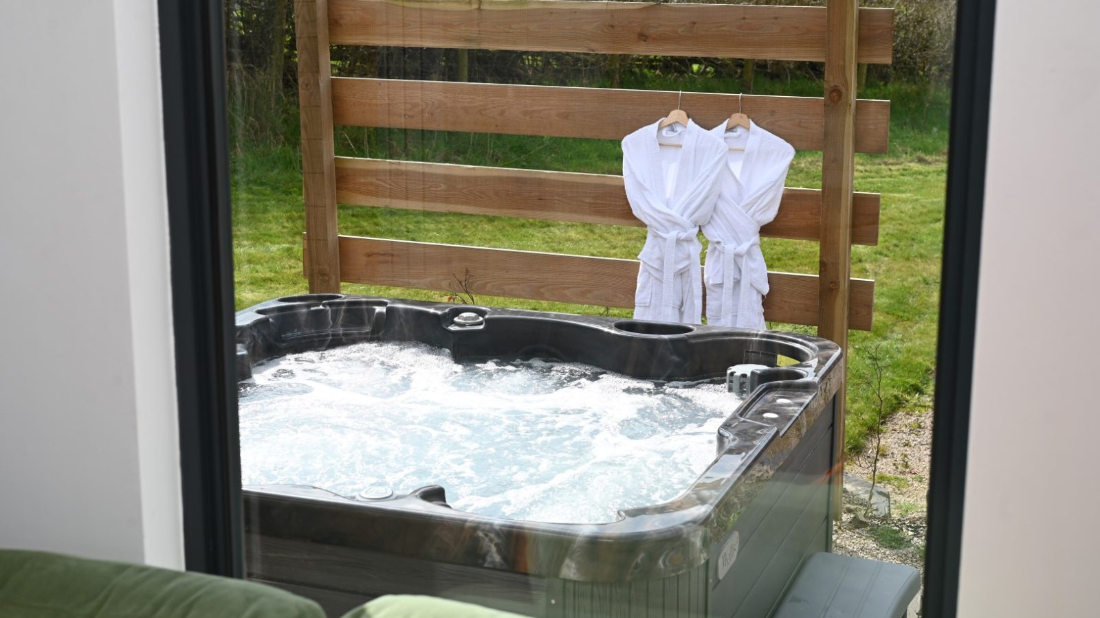 Peering through the window, discover Belan View's bubbling outdoor hot tub, where two white bathrobes hang on a wooden fence in the grassy yard. This serene sanctuary offers an inviting relaxation spot at one of the finest lodges around.