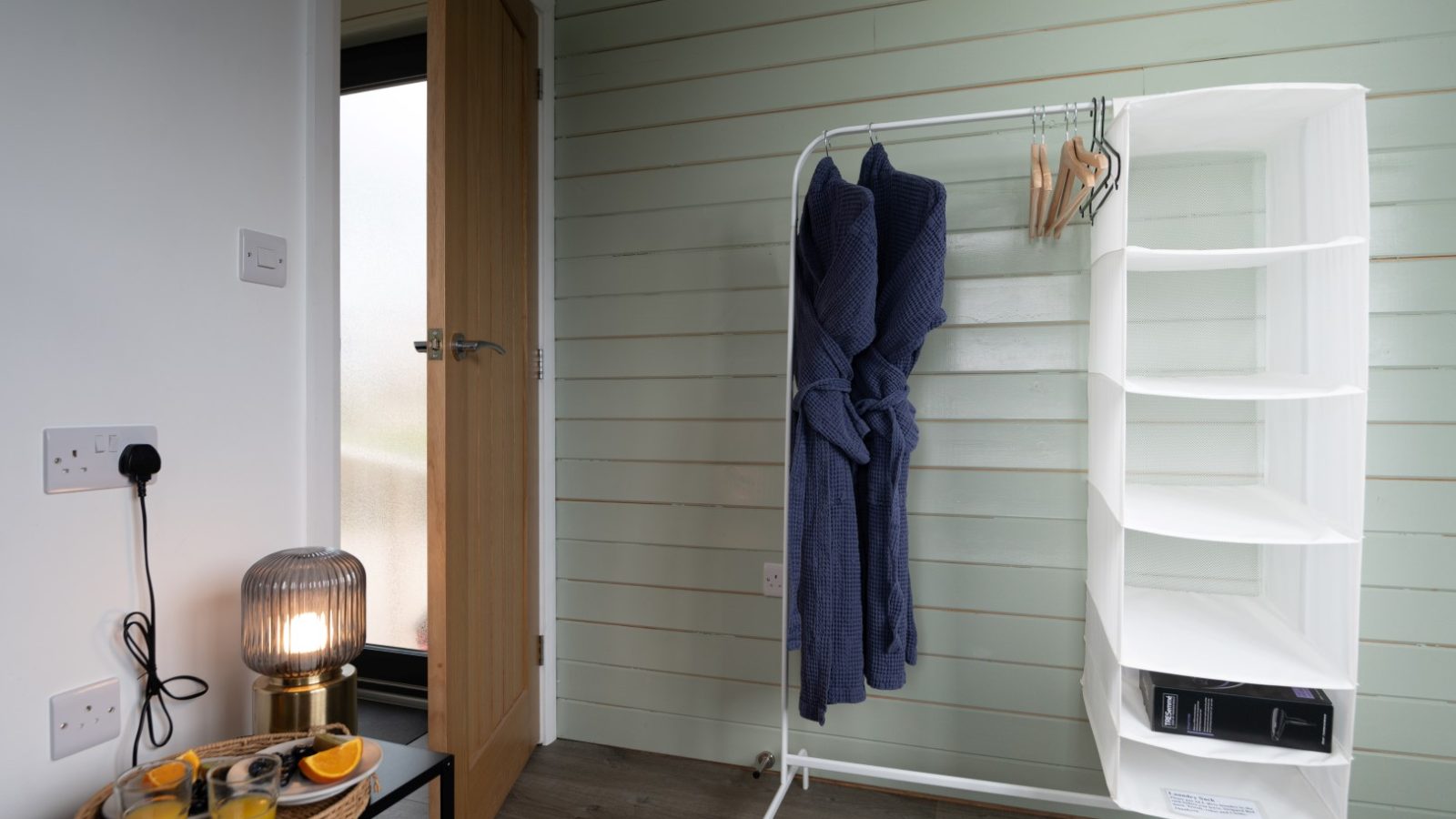 A cozy room in Belan View boasts a wooden floor and features a garment rack with two blue robes. Nearby, a shelf holds a black folder. A small table with juice, oranges, and a lamp sits beside the door and frosted window—capturing the essence of The Lodges beautifully.