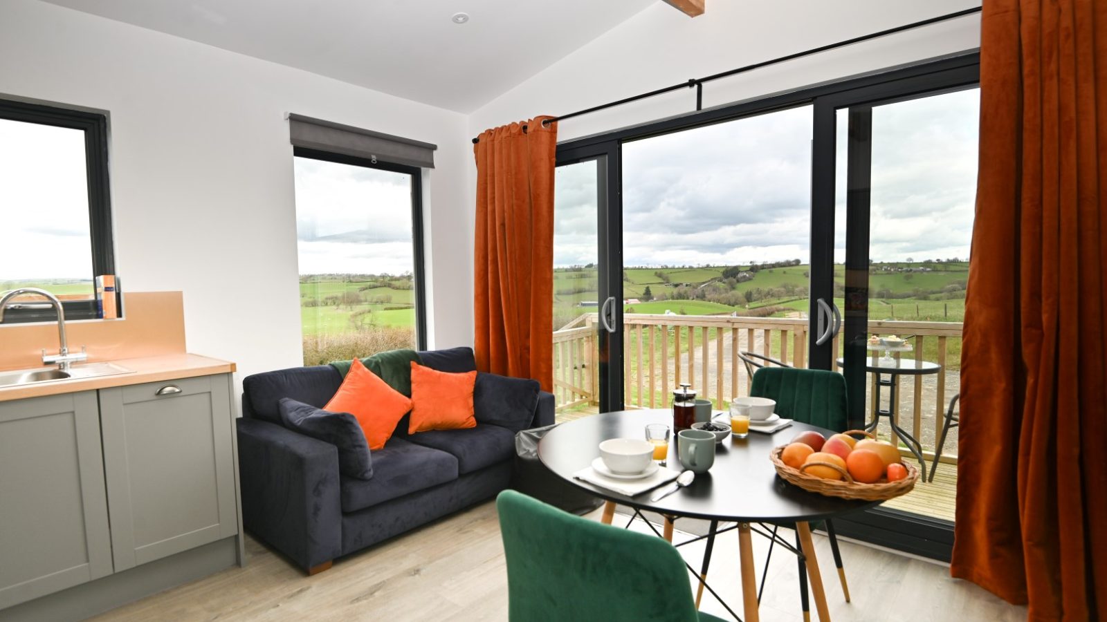 A cozy living room at Belan View Lodges features a gray sofa with orange and green cushions, and a round dining table set for breakfast. Large windows with orange curtains reveal a scenic vista of green hills beneath a cloudy sky.