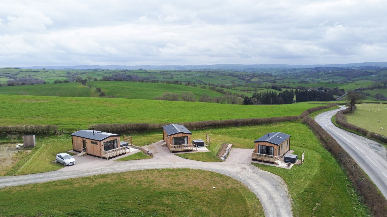 Three charming lodges, known as Belan View, are nestled in an open, grassy landscape with rolling hills in the background. A paved road winds alongside the property, and a cloudy sky extends overhead.