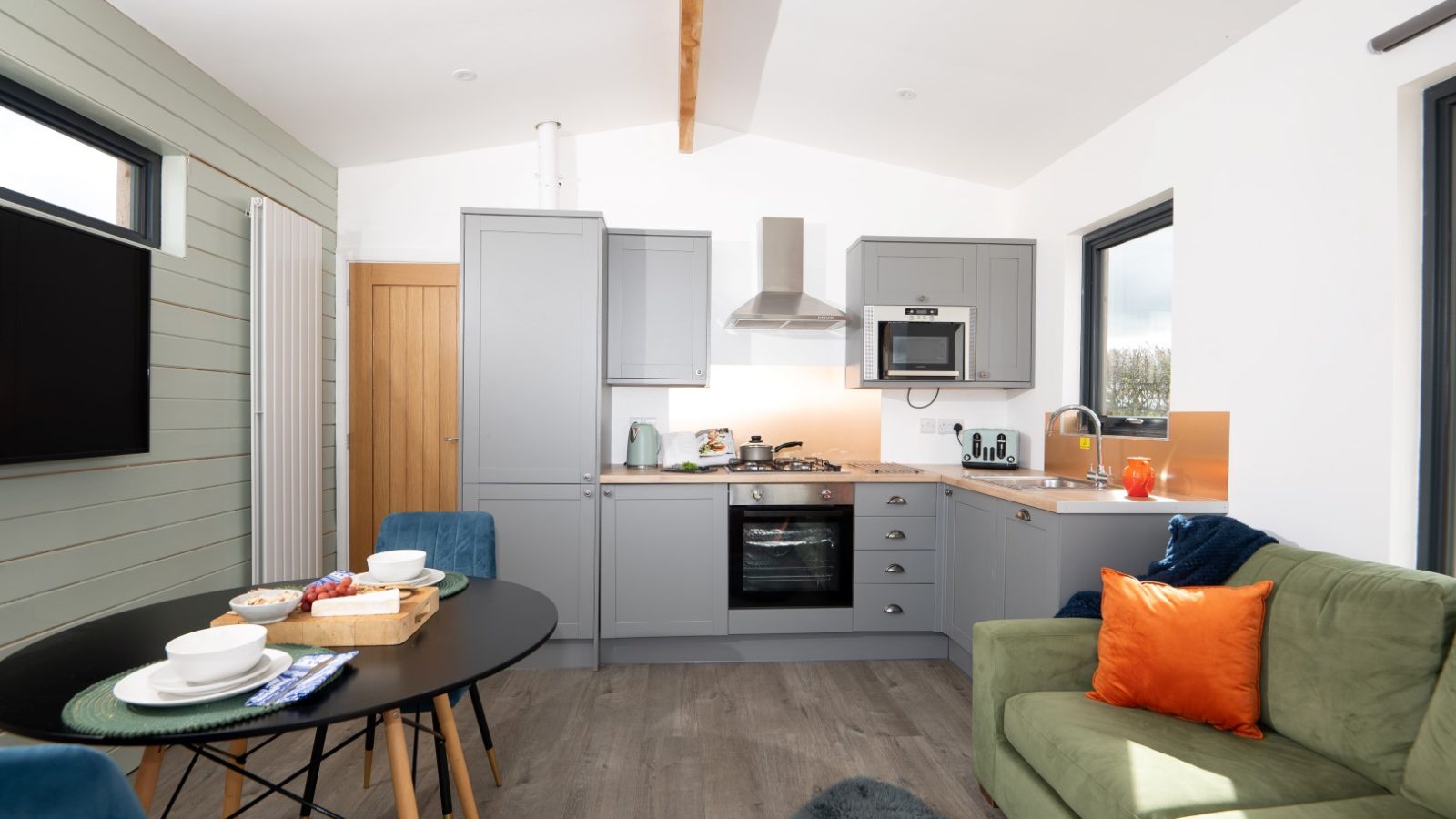 The modern kitchen and living area at Belan View features gray cabinets, an oven, and a microwave. A round dining table with dishes complements the green sofa adorned with orange pillows. Sunlight floods through large windows, highlighting the cozy, contemporary decor.