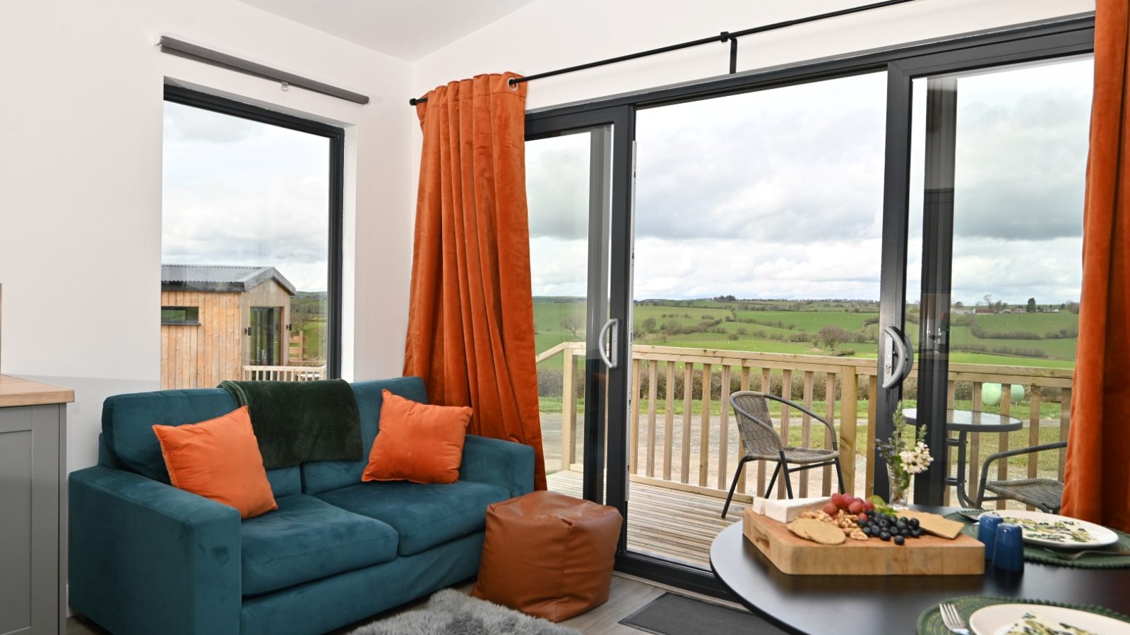 A cozy living room in Belan View features a teal sofa adorned with orange pillows and a plush rug. Large sliding glass doors open to a wooden deck, offering a scenic view of the green landscape. In the foreground, a table with a cheese and fruit platter completes this inviting scene at The Lodges.