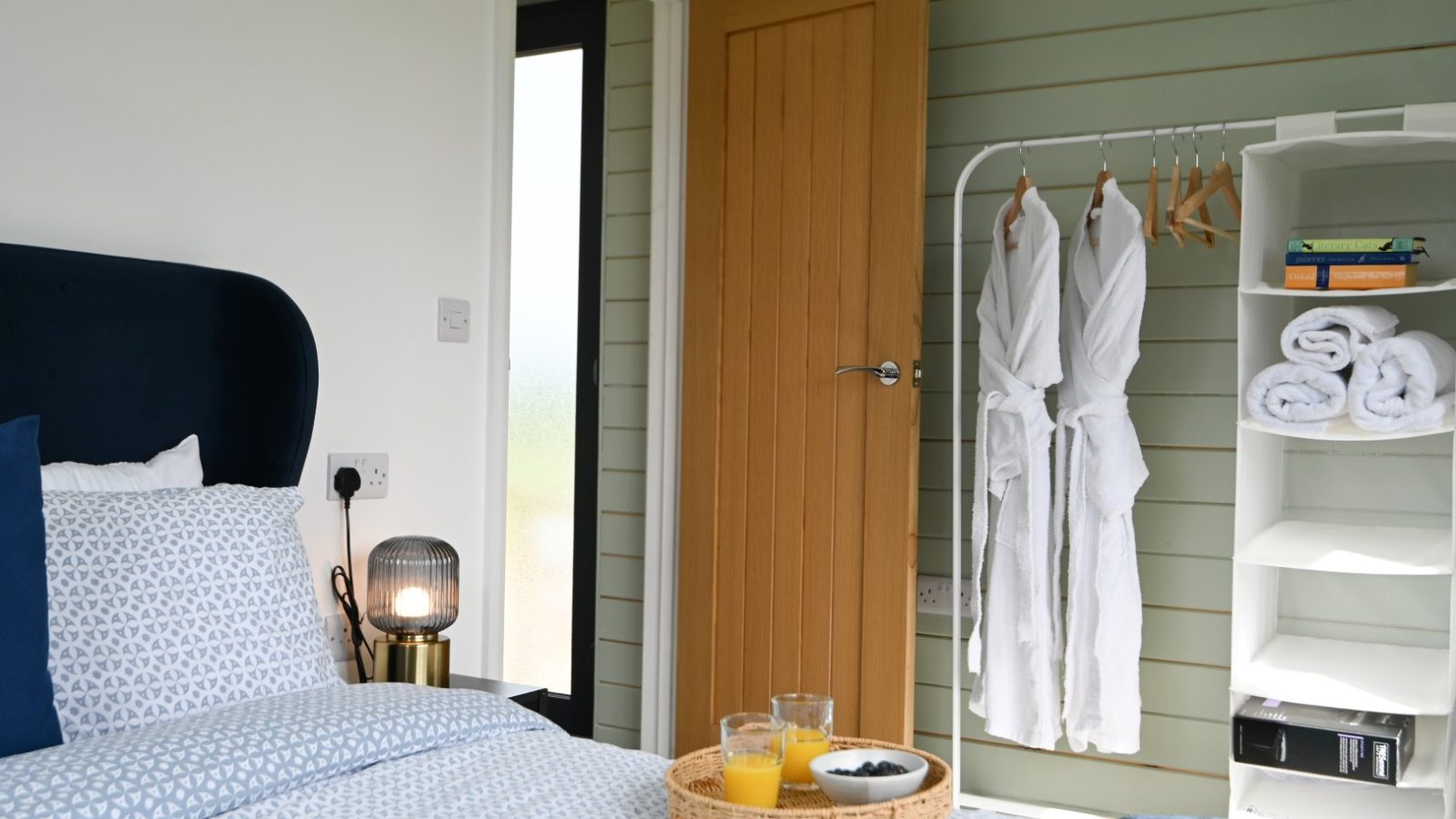 Cozy bedroom at Belan View Lodge with a neatly made bed, patterned pillows, and a bedside lamp. A tray with juice and snacks awaits. A wooden door is slightly open, revealing a rack with robes and shelves of towels and books against the pastel green shiplap wall.