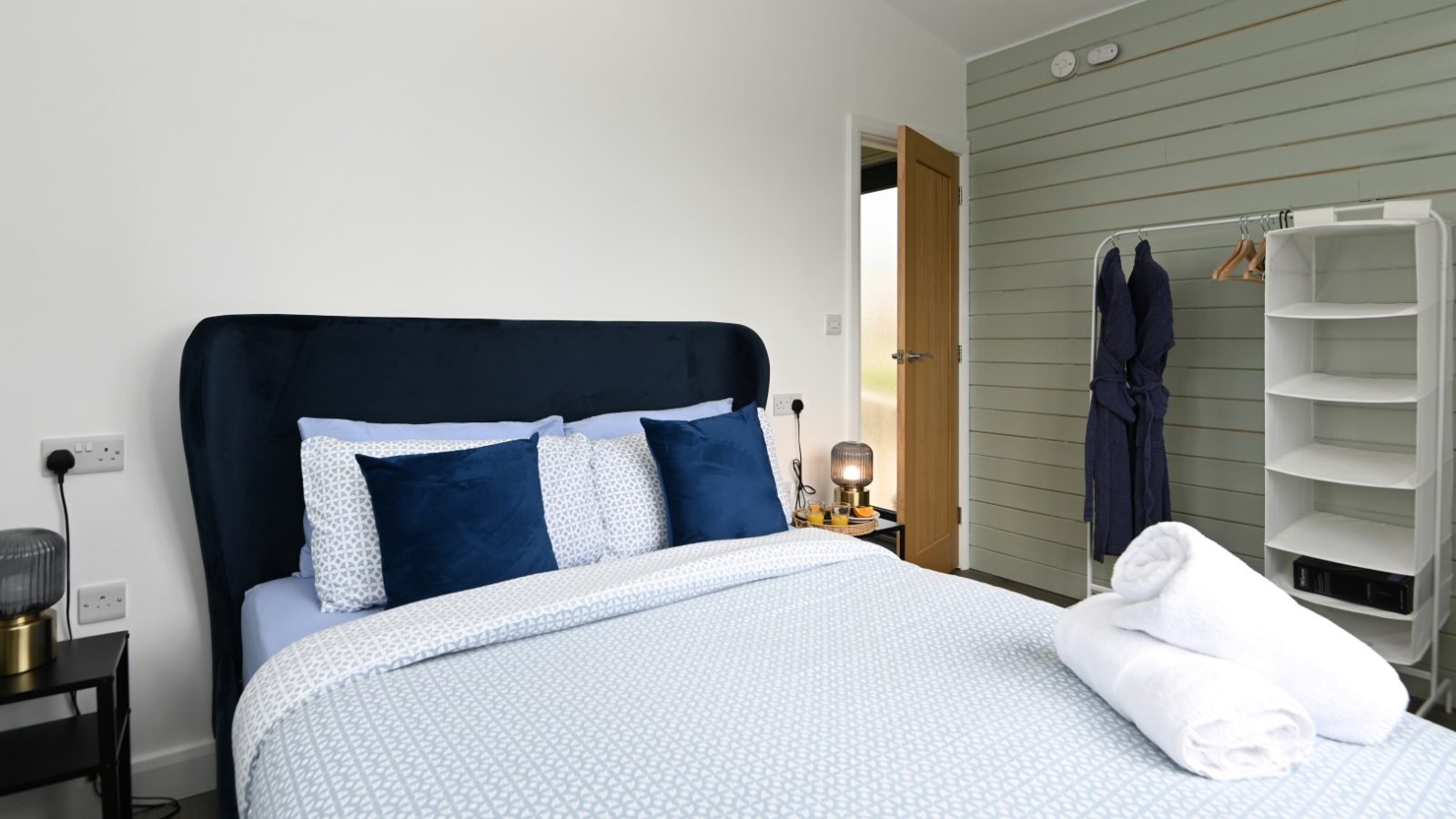 A modern bedroom at Belan View features a neatly made bed with blue and white bedding, flanked by two small bedside tables. One table holds a lamp. A wooden door is slightly ajar, while a white open closet with a robe hangs invitingly on the right.